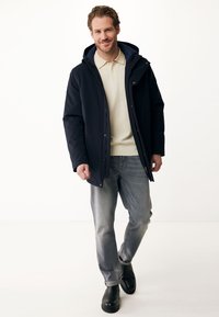 Veste imperméable bleu marine avec capuche, fermeture éclair à l'avant et boutons pressions, associée à un polo beige clair et un jeans gris. Des bottines noires à la cheville complètent le look.