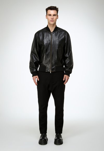 Fiatal férfi áll, fekete bőr bombázó dzsekit, fekete nadrágot és fekete masszív csizmát visel, egy egyszerű világos háttér előtt.