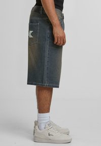 Denim shorts in a faded blue shade, knee-length with a back pocket featuring a large embroidered "X" logo. Worn with white sneakers.