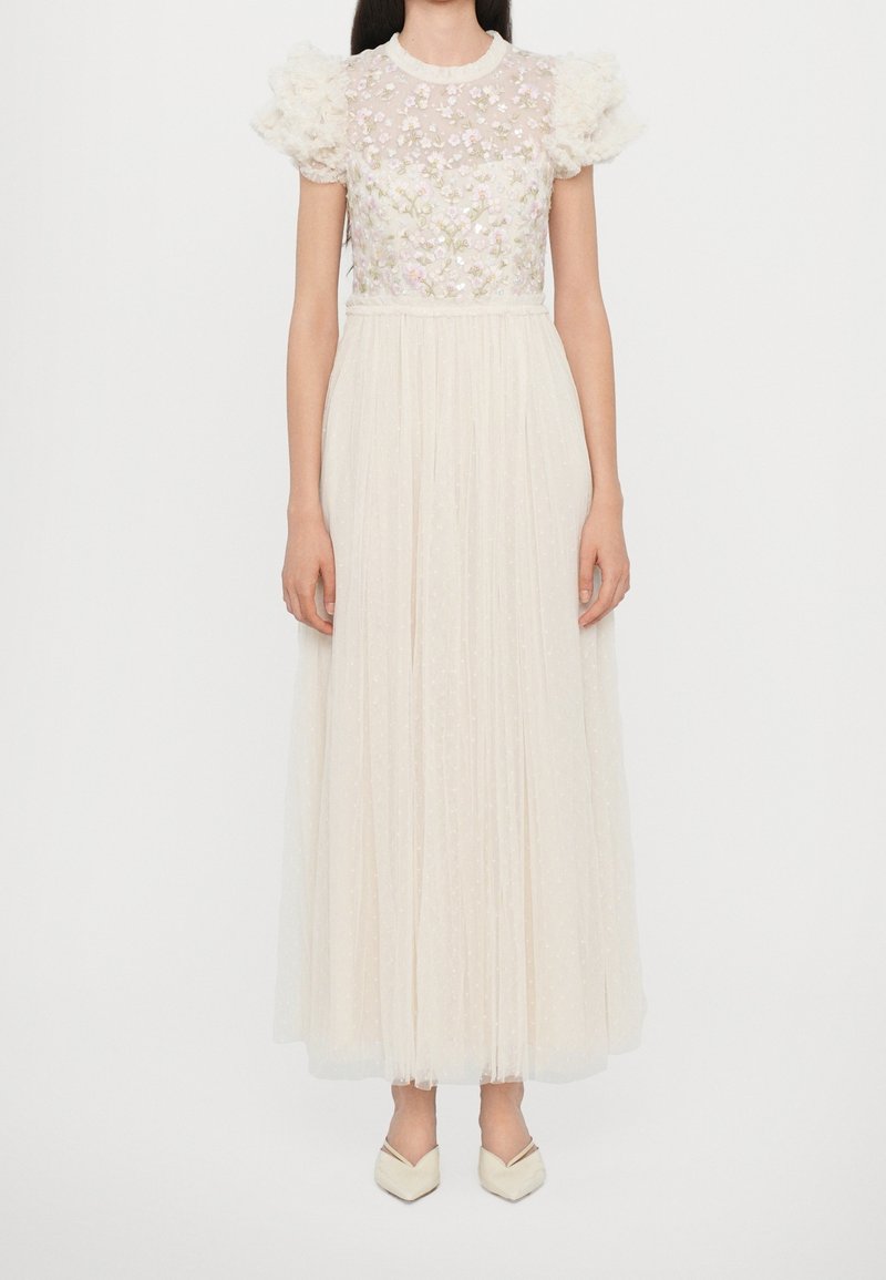 Femme portant une robe crème longue jusqu'au sol avec des manches bouffantes et une broderie florale sur le corsage transparent, debout devant un fond blanc.