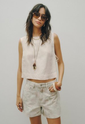 Ung kvinde med langt mørkt hår bærer solbriller, en ærmeløs beige top, lyse mønstrede shorts, vedhængshalskæde og chunky armbånd.