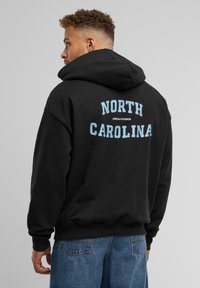 Zwarte hoodie van dik materiaal. Heeft "NORTH CAROLINA" in lichtblauwe letters op de achterkant. De voorkant is eenvoudig. Casual pasvorm.