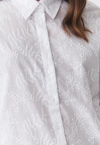 Blouse blanche brodée avec un motif de feuilles, un col et des boutons, fabriquée en tissu léger avec une finition douce et texturée.
