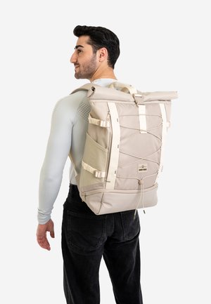 Beige roll-top backpack with adjustable straps, front zippers, and a textured surface. Designed with a unique elastic closure and side pockets.