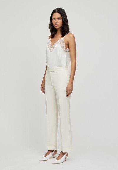 Camicetta in satin di seta con rifiniture in pizzo, abbinata a pantaloni cream a vita alta e suole bianche appuntite. Design semplice ed elegante.
