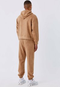Beige fleece hoodie en joggingset gemaakt van zachte, gestructureerde stof, met een capuchon en elastische manchetten, gecombineerd met witte sneakers.