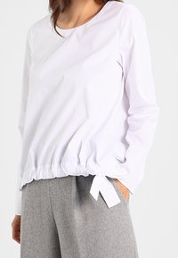 Haut blanc à manches longues avec encolure arrondie, taille élastiquée et détail de nœud sur le côté, associé à un pantalon gris. Texture de tissu lisse.