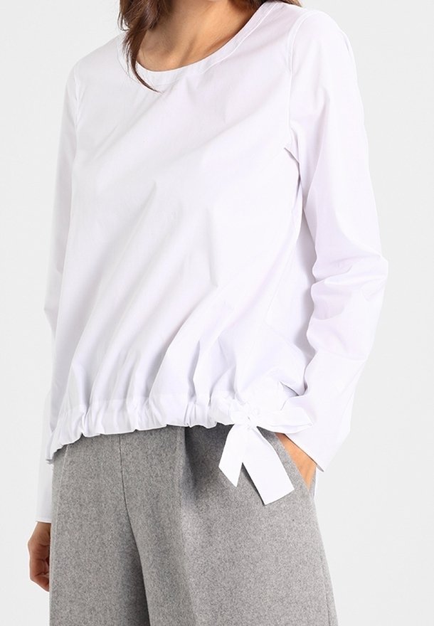 Haut blanc à manches longues avec encolure arrondie, taille élastiquée et détail de nœud sur le côté, associé à un pantalon gris. Texture de tissu lisse.