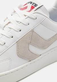 Białe skórzane sneakersy z jasnobeżowym zamszowym akcentem w postaci logo, białymi sznurowadłami oraz czerwoną i białą metką z logo na języku.