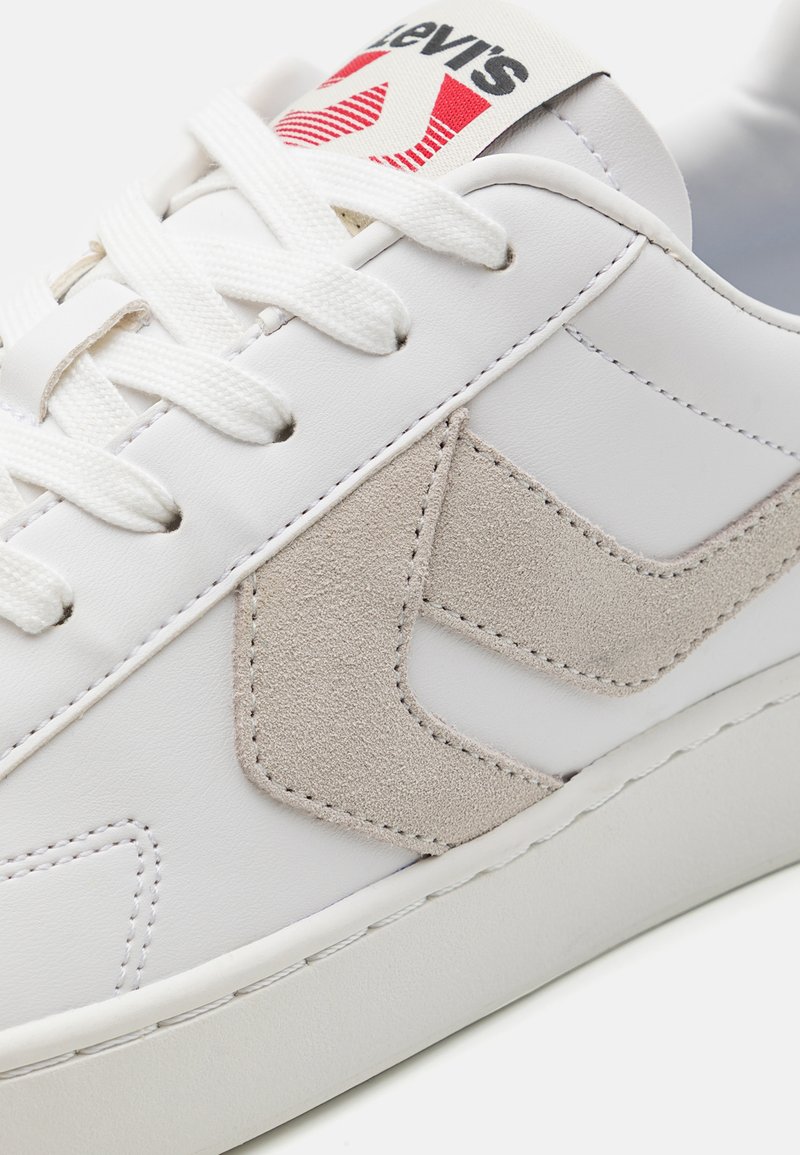 Baskets en cuir blanc avec un accent en suède beige clair sur le logo, lacets blancs et un onglet de marque rouge et blanc sur la languette.