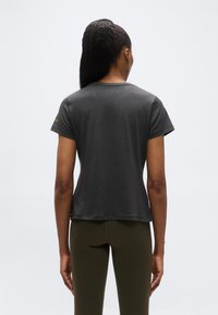 Γυναίκα με σκούρα αλογοουρά που φοράει κοντομάνικο σκούρο γκρι μπλουζάκι και ελιά πράσινο κολάν, έχει την πλάτη της στραμμένη προς ένα απλό φωτεινό φόντο.