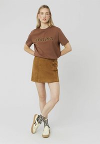 T-shirt marron avec le mot « FEELING » en police à motifs, associée à une jupe en daim beige avec deux détails de boutons et des baskets blanches portées avec des chaussettes texturées.