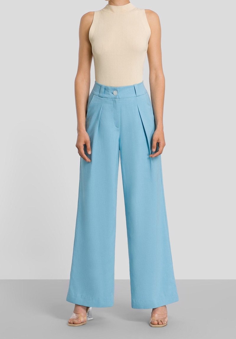 Pantalon bleu clair à jambes larges avec plis, taille haute et fermeture par un seul bouton, associé à un haut sans manches beige côtelé. Texture claire et lisse.