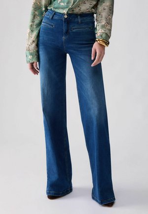 Kvinde iført blå jeans med høj talje og vide ben samt en blomsterprintet grøn bluse med guldbøjler på højre håndled mod hvid baggrund.