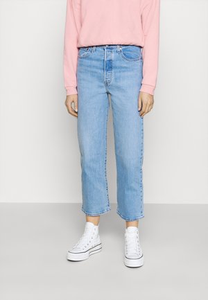 Personne portant un jean droit court bleu clair, des baskets montantes blanches et un haut à manches longues rose, debout sur un fond uni.