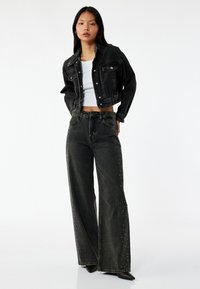 Sort denim cropped jakke og vide sorte jeans med nitter langs kanten. Modellen bærer en hvid top og sorte hæle.