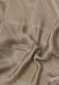 Beige kuvioitu huivi, joka on valmistettu pehmeästä kankaasta ja jossa on teksturoitu pinta valkoisilla abstrakteilla kuvioilla sekä fringed-reunoilla.