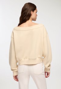 Felpa beige oversize con design off-shoulder, polsini a coste e orlo a coste. Tessuto morbido con una vestibilità comoda, abbinata a pantaloni di colore chiaro.