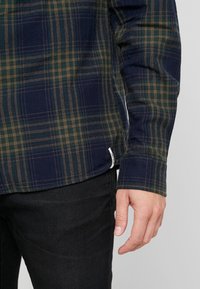 Oikea käsi ja käsi henkilöstä, joka käyttää vihreää ja merivihreää ruudullista pitkähihaista paitaa ja mustia housuja yksinkertaista taustaa vasten.