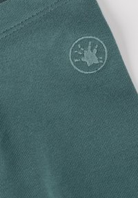 Grüner Baumwollstoff mit einer weichen, strukturierten Oberfläche. Enthält ein rundes gesticktes Logo, das eine Pfote darstellt.