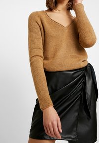 Pull en tricot brun clair avec un col en V et des manches longues, associé à une jupe portefeuille en simili cuir noir avec un détail de nouage.