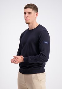 Granatowy dres z bawełnianej tkaniny, charakteryzujący się okrągłym dekoltem, prążkowanymi mankietami oraz małą naszywką z logotypem na lewym rękawie.