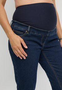 Trudnički traperice s tamnoplavim denim finišem, s rastezljivim mornarskim trakom za trbuh, petljama za remen i kontrastnim narančastim šavovima.