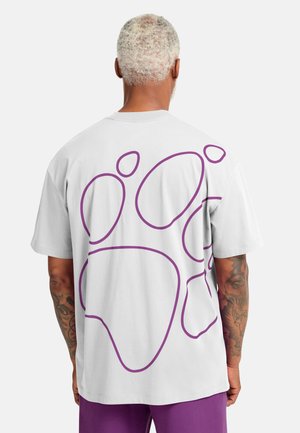 Maglietta bianca con un grande disegno di un'impronta di zampa viola sul retro. Maniche corte e vestibilità comoda. Il tessuto appare liscio e morbido.