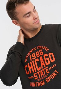Felpa nera con una grafica audace rossa, testo "1986 CHICAGO STATE" e "VINTAGE SPORT". Collo e polsini a costine, texture morbida.