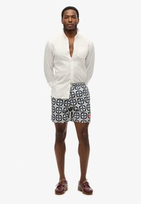 Camisa de lino blanca, pantalones cortos de baño azul marino con diseño floral y zapatos burdeos. Los detalles notables incluyen un pequeño bolsillo en la camisa.