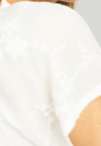 Weiße Bluse mit floralem Stickerei, kurzen Ärmeln und einem leichten, strukturierten Stoff. Der Ausschnitt ist rund und weich.