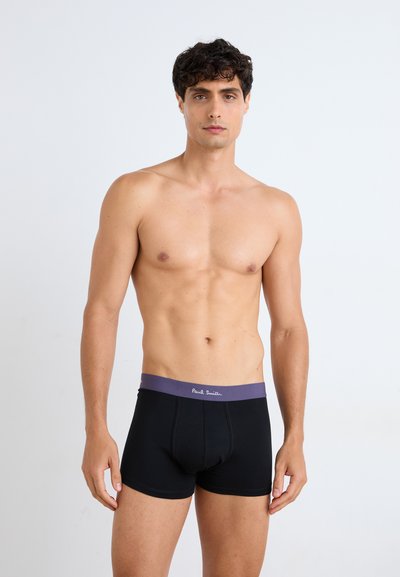 Sorte bokserbriefs med en lilla midjebånd, med synlig merkevare. Stoffet ser glatt ut, former seg til kroppen og strekker seg.