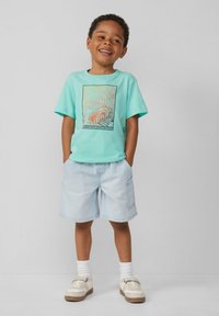 Hellblaues Baumwoll-T-Shirt mit Palmenblatt-Graphic, kombiniert mit hellen Jeansshorts. Die Schuhe sind weiße Sneaker mit braunen Sohlen.