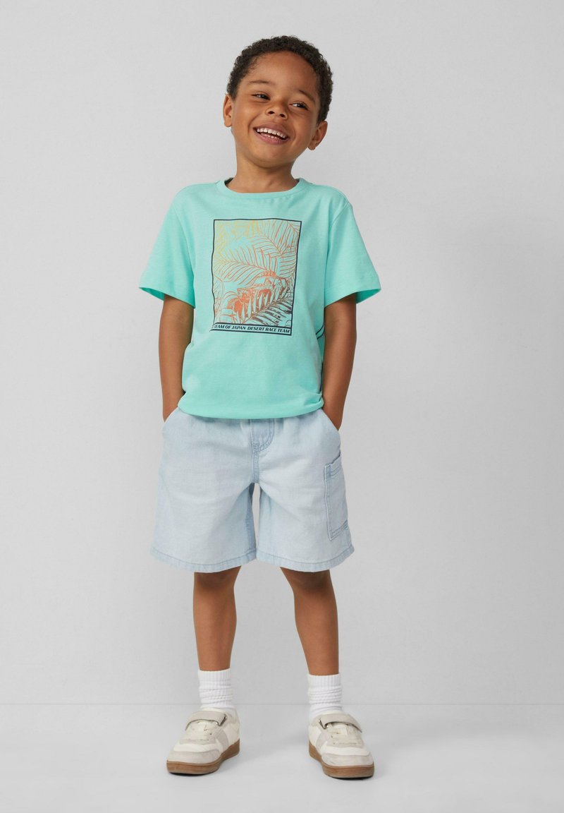 Hellblaues Baumwoll-T-Shirt mit Palmenblatt-Graphic, kombiniert mit hellen Jeansshorts. Die Schuhe sind weiße Sneaker mit braunen Sohlen.