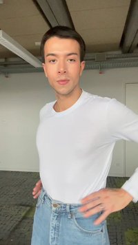 Young man with short dark hair wearing a white long-sleeve shirt and blue jeans, standing indoors with industrial ceiling visible.