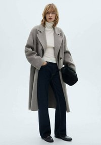 Manteau en laine gris clair avec un design croisé, porté sur un col roulé blanc et un jean foncé. Un sac à main noir et des chaussures plates complètent le look.