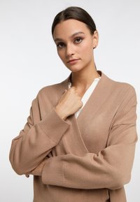 Beige wikkelcardigan met een diepe V-hals en brede mouwen. Zachte gebreide textuur, geribbelde manchetten, gedragen over een witte top.