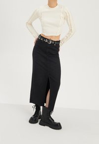 Frau in einem cremefarbenen langen Ärmeln Cropped-Top, schwarzem, gebändertem Midisch skirt mit einem Schlitz vorne, Netzstrumpfhosen und schwarzen Schnür-Stiefeln steht.