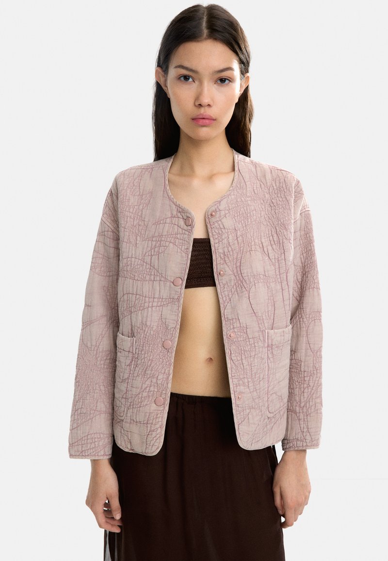 Jasnoróżowa kurtka zapinana na guziki z teksturowanym wzorem, okrągłym dekoltem i dwiema przednimi kieszeniami. Materiał wydaje się miękki i pikowany.