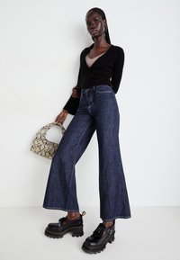 Jeans a gamba larga blu scuro con cuciture bianche a contrasto, abbinati a un maglione avvolgente nero e a scarpe platform nere chunky. Borsa con stampa serpente.