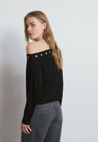 Joven mujer con cabello largo y rubio, vestida con un top negro de hombros descubiertos con ojales metálicos y jeans gris oscuro, mirando por encima del hombro.