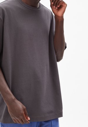 Chemise oversize à manches courtes de couleur gris foncé portée par une personne, associée à un pantalon bleu, une main près du visage et l'autre détendue.