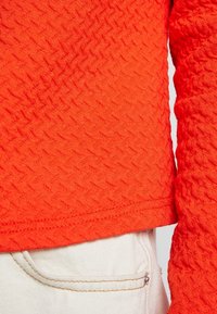 Oranssi tekstuurinen neulepaita, jossa on timanttimäinen kuvio, pitkät hihat ja helma, joka päättyy juuri vyötärön ylle, yhdistettynä valkoisiin housuihin.