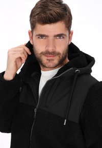 Chaqueta negra de forro polar con una sección superior acolchada, cierre de cremallera y capucha ajustable. Presenta una textura suave y un corte holgado.