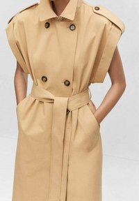 Beżowy płaszcz trenczowy z podwójnym zapięciem, poszerzonymi ramionami, dużymi guzikami i pasującym paskiem. Posiada boczne kieszenie i gładką fakturę.