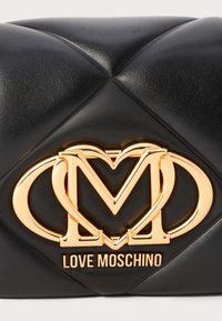 Melnā quiltotā ādas rokassoma ar zelta tono "M" logotipu sirdshape formā. Apakšējā tekstā ir rakstīts "LOVE MOSCHINO."