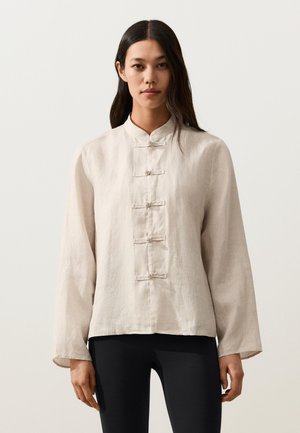 Woman wearing a beige long-sleeve linen top with five knot buttons down the front and black pants, standing against a plain background.