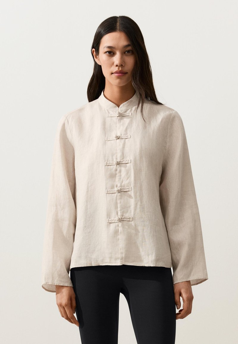 Femme portant un haut en lin beige à manches longues avec cinq boutons en forme de nœud sur le devant et un pantalon noir, debout devant un fond uni.