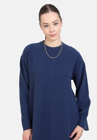Modrá oversized šaty vyrobené z měkké látky, s dlouhými rukávy a kulatým výstřihem. Obsahují také jednoduchý stříbrný řetízek.