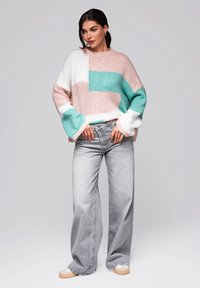 Yli suuri vaaleanpunainen, teal- ja valkoinen neule, jossa on neulottu rakenne, yhdistettynä korkeavyötäröisiin vaaleanharmaisiin farkkuihin ja tasapohjaisiin rentoihin kenkiin.